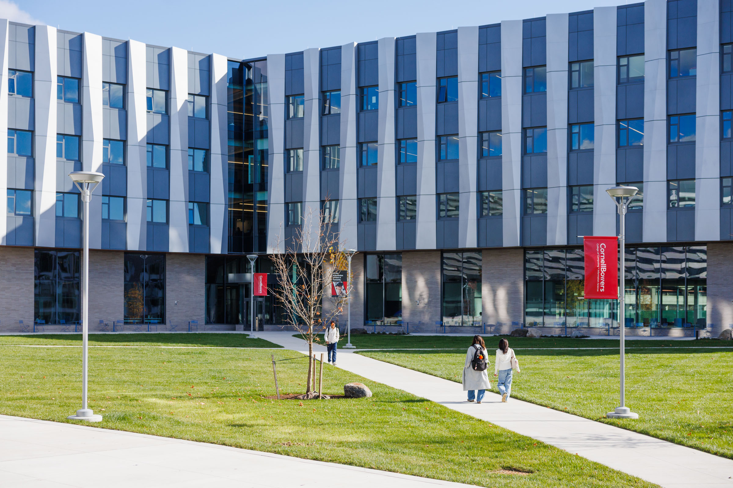 students walk along a paved path surrounded by green grass in front of a gray building