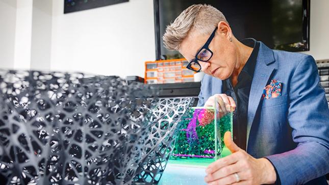 A white woman with short styled hair, dark rimmed glasses, and a blazer lookin at a 3d printed model.