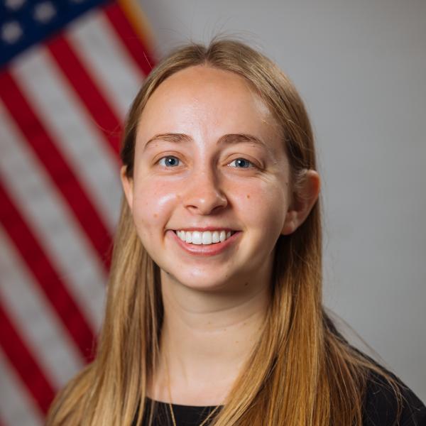 a female student with long blonde hair and a long sleeve black shirt sits in front of an American flag