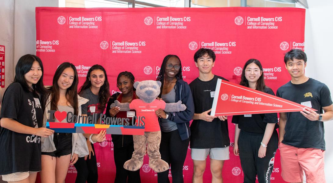 A diverse group of 8 college students pose with Cornell Bowers banners and flags.