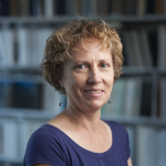 A color photo of woman with short curly hair. Books in background.