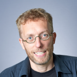 A photo of Kilian Weinberger, a man with curly blond hair, glasses, a beard and mustache, in a gray shirt in front of a gray background