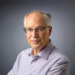 A photo of Bart Selman, a man with white hair and glasses in a lavender shirt in front of a gray background