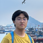 A color photo of a man with glasses in front a bay with boats.