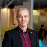 A portrait of Thorsten Joachims, a man with gray hair and a dark red shirt smiling at the camera