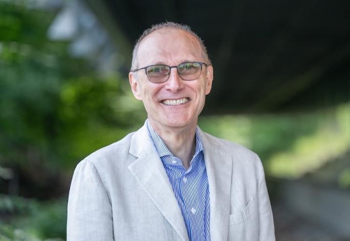 Man with glasses, short hair, blue shirt and tan jacket smiling at the camera