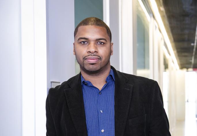 A photo of Hakim Weatherspoon, a mwan with buzzed black hair and beard and mustache, in a blue shirt and black jacket, in a hallway