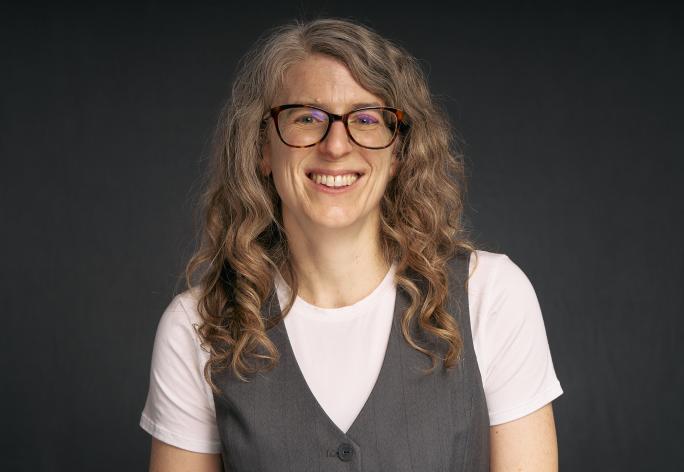 A color photo of a woman with long curly brown hair and glasses in front of a gray background