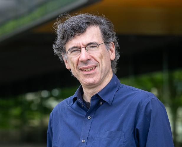 A portrait of Jon Kleinberg, a man with curly gray hair, glasses and a blue shirt, in front of Gates Hall