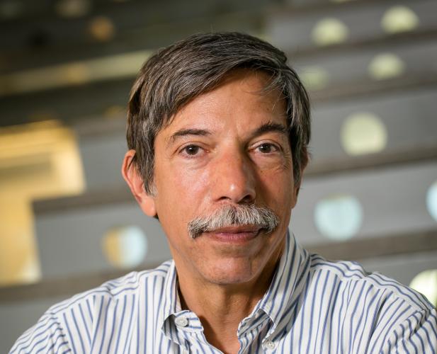 A photo of Fred Schneider, a man with dark gray hair and a mustache, in a striped shirt in front of a staircase