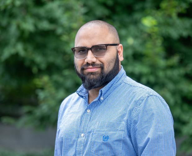 A photo of Sainyam Galhotra, a man with a shaved head, a dark beard, dark glasses and a blue shirt in front of a leafy background.