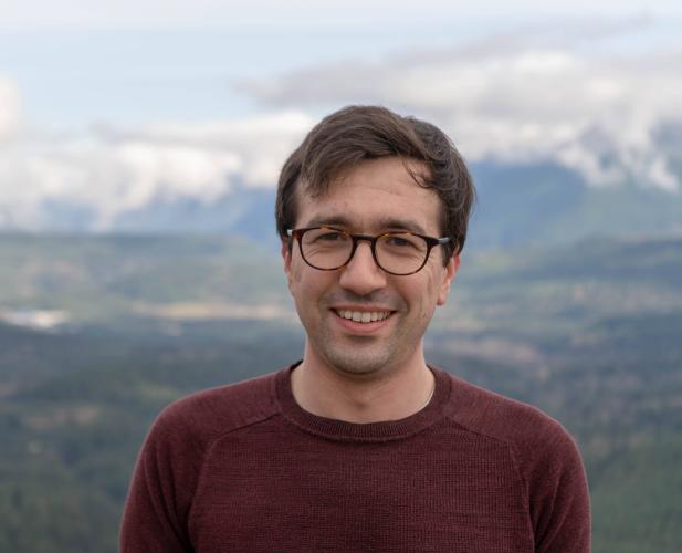 A photo of Nicholas Spooner, a smiling man with brown hair and glasses in a maroon sweater, in front of a mountain range.