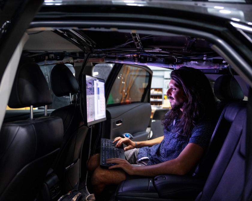 inside of a dark car a man with long dark hair sits in the backset looking at a computer screen