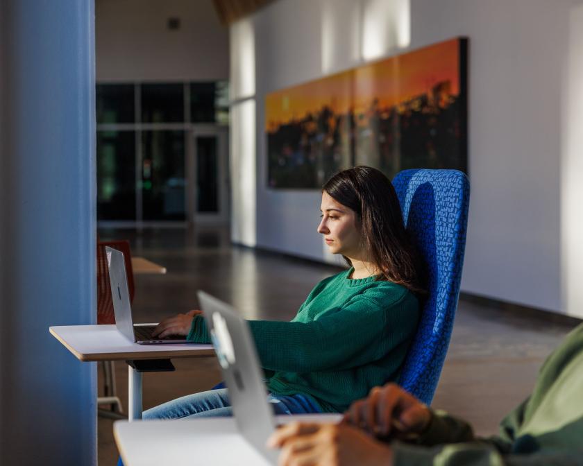 a female student with brown hair sits and works on her laptop in the hallway